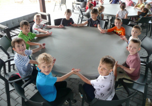 dzieci siedzą przy okrągłych stołach i trzymają się za ręce