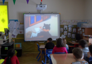 dzieci siedząc przy stoliczkach oglądają film na tablicy interaktywnej dotyczący tematyki zajęć