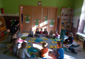 dzieci siedząc w kole uczestniczą w zajęciach logorytmicznych, jedno z dzieci odpowiada na pytania