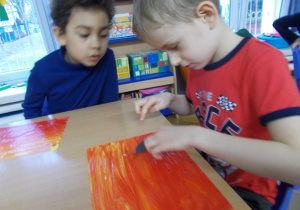 Juniorzy przy stolikach wykonują pracę plastyczną: malowanie farbami plakatowymi