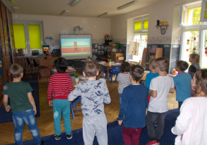 Juniorzy w swojej sali wykonują ćwiczenia gimnastyczne pokazane na tablicy interaktywnej