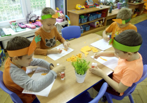 Juniorzy przy stoliczkach z emblematami marchewki na głowie wykonują pracę plastyczną