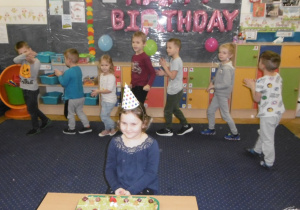 Juniorzy w kole na dywanie śpiewaja piosenkę urodzinową, dziewczynka w czapeczce urodzionowej przed tortem