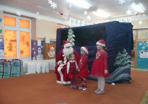 trzy dziewczynki ubrane na czerwono śpiewają kolędę Mikołajowi