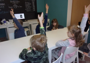 Juniorzy przy stole w bibliotece podnoszą ręce chcąc odpowiedzieć na pytanie