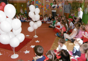 dzieci siadzą na sali gimnastycznej, w tle białe balony na stojakach