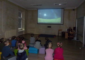 Juniorzy w auli Placentarium sidzą na ławeczkach i oglądają prezentację z projektora