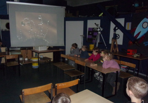 Juniorzy siedzą w ławeczkach i oglądają film z projektora