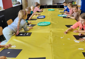 dzieci przy stole przykrytym żółtą folią układają na czarnych kartonach kawałki białego papieru