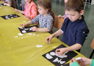 dzieci przy stole przykrytym żółtą folią układają na czarnych kartonach kawałki białego papieru