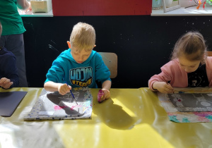 troje dzieci p[rzy stole maluje farbami plakatowymi