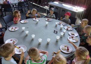 dzieci przy okrągłych stołach jedzą kiełbaski z grilla