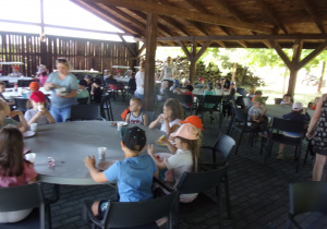 dzieci przy okrągłych stołach jedzą kiełbaski z grilla