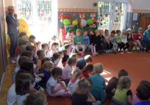 dzieci na sali gimnastycznej podczas słuchania wierszy czytanych przez pracownikó przedszkola