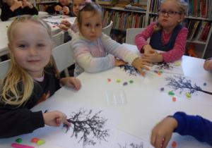 dziewczynki przy stoliku przyklejają z plasteliny liście na sylwecie drzewa