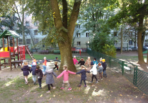Juniorzy w kole tańczą dookoła drzewa w ogrodzie przedszkolnym