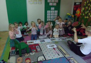 Tuptusie siedzą na dywanie prze ilustracjami i naśladują nauczyciela